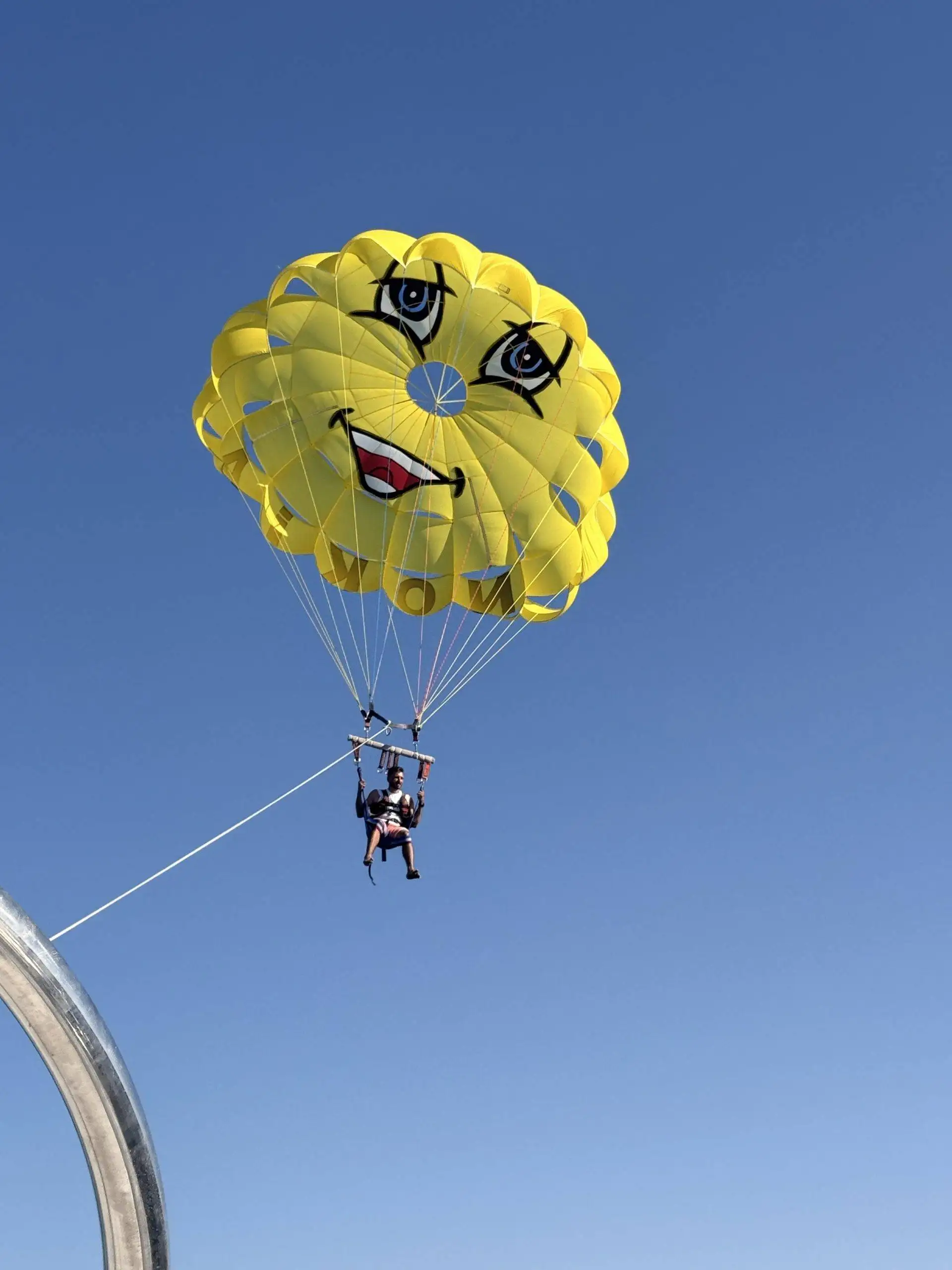 Person parasailing in a clear blue sky under a bright yellow parachute with a smiling cartoon face