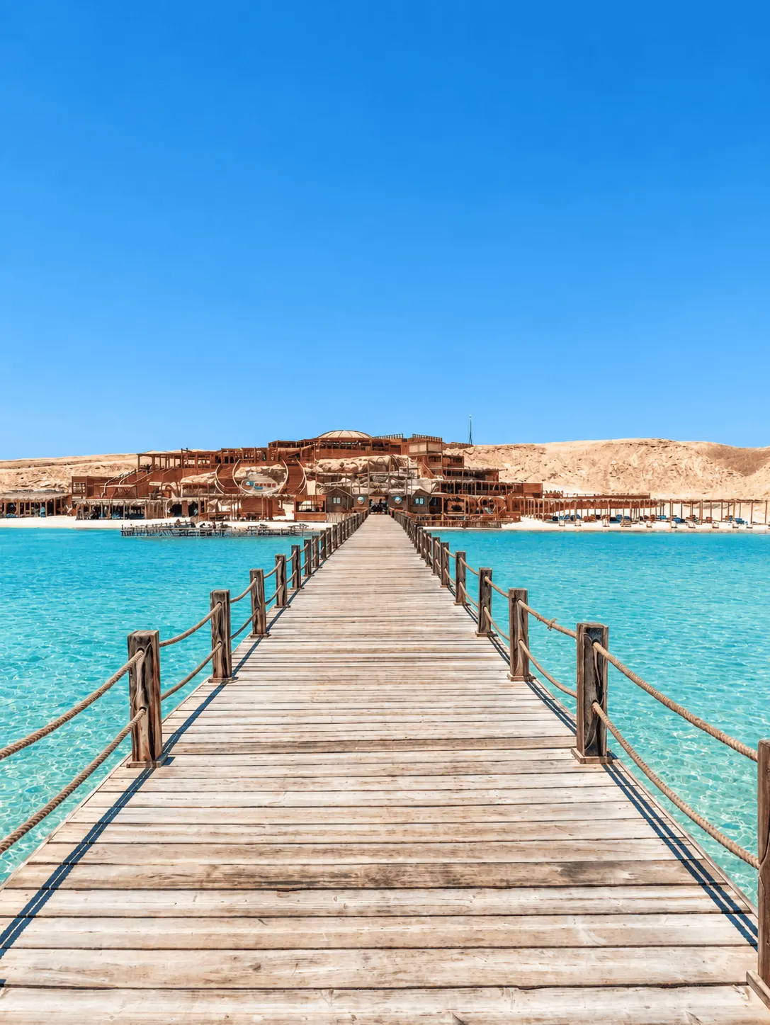 A wooden pier extending across clear turquoise water toward a desert beach resort set against sand dunes under a bright blue sky