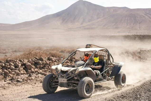 Two people wearing face bandanas and sunglasses driving an off-road buggy along a dusty desert track with arid hills behind